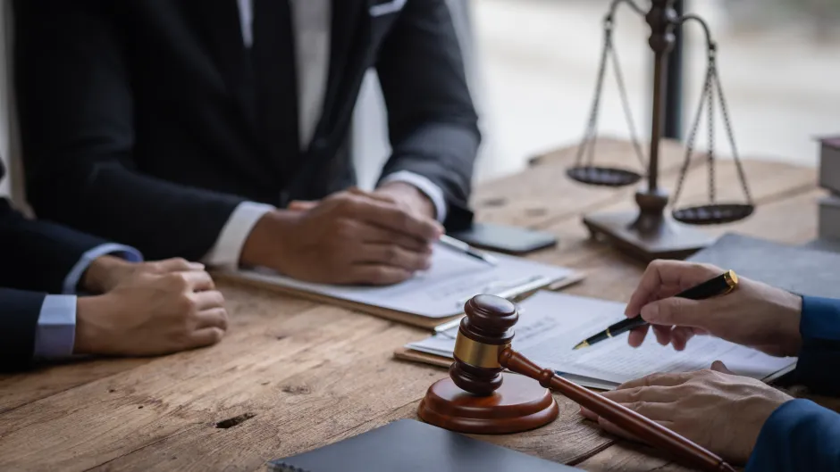 Blick auf einen Schreibtisch, auf dem Verträge liegen, im Hintergrund Justizias Waage